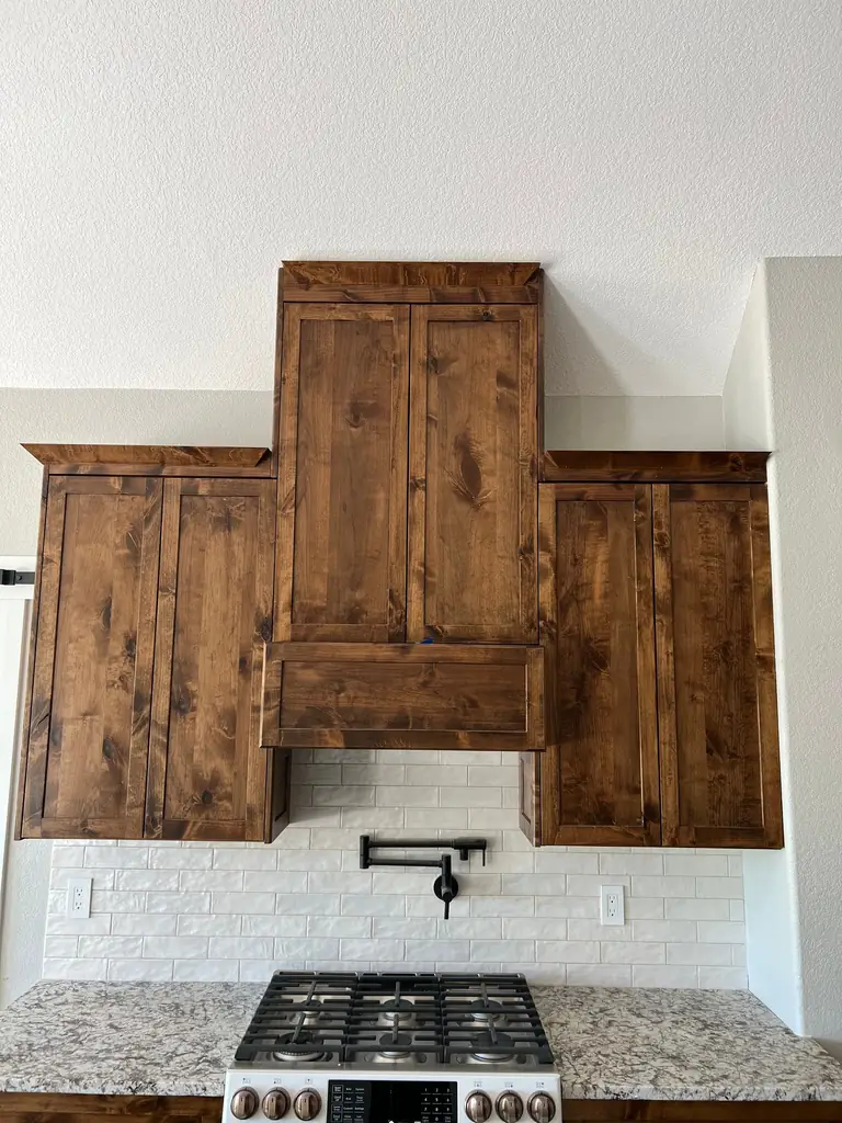 Alder range hood surround and matching wall cabinets over a brick-look backsplash and pot filler