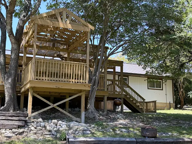Elevated wood deck with a gabled pergola, railings and stairs in a tree-shaded yard
