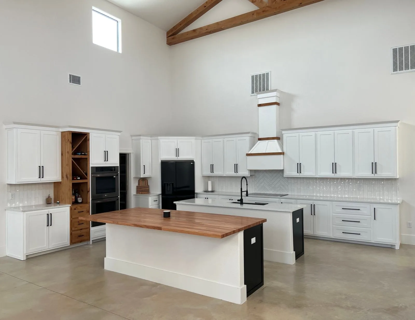 Vaulted white shaker kitchen with exposed beams, two islands, a wood-banded hood and a wine cabinet