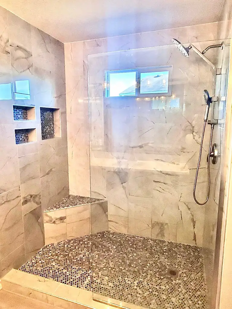 Walk-in shower with marble-look tile, recessed niches, a tiled bench and pebble floor behind glass